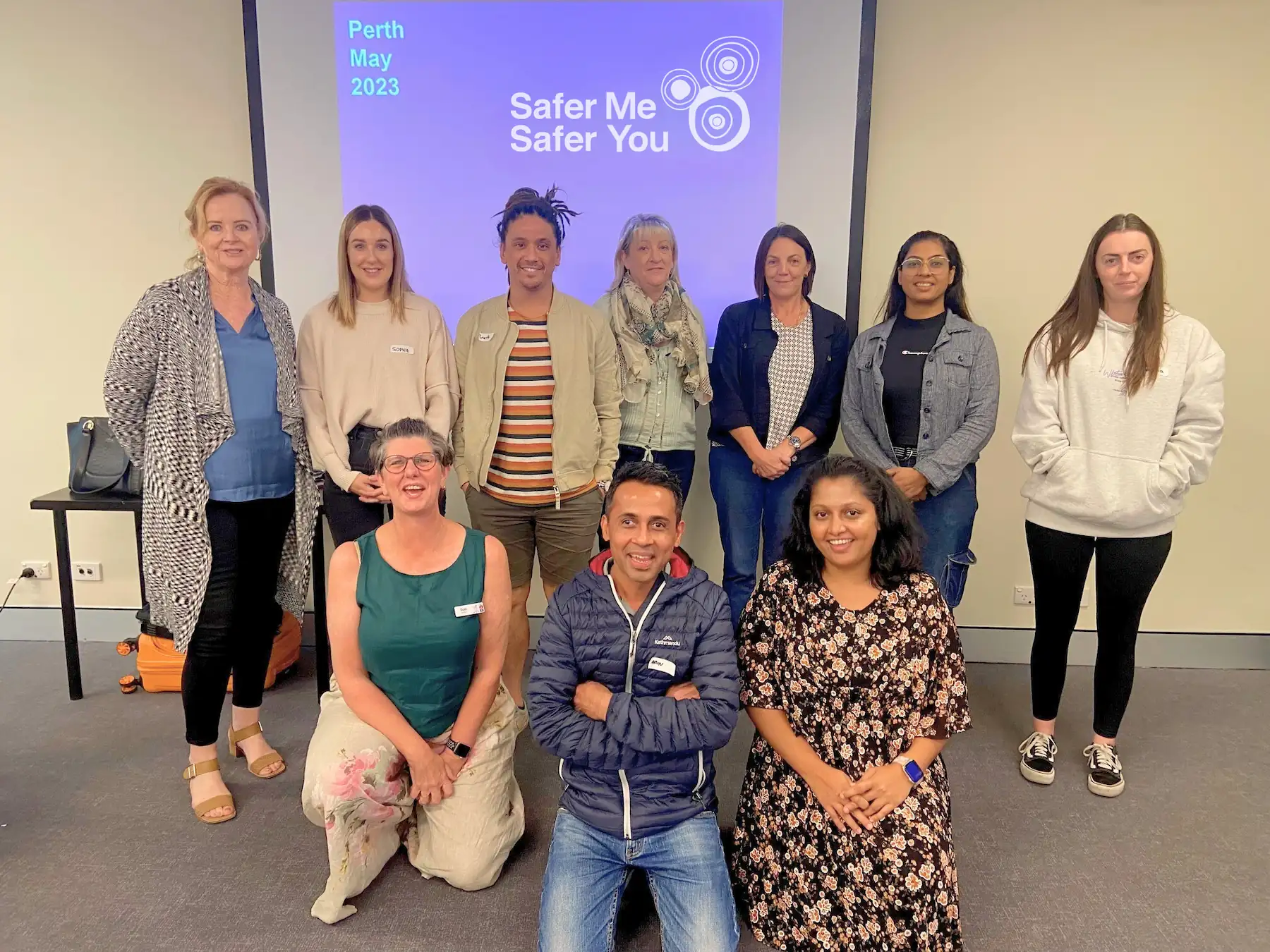 11 people at a 'Safer Me Safer You' event in Perth, May 2023. They are standing and kneeling in front of a projector screen displaying the event name.
